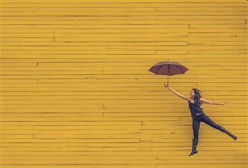 Young woman jumping gleefully with an umbrella against a bright yellow wall. Photo by Edu Lauton.